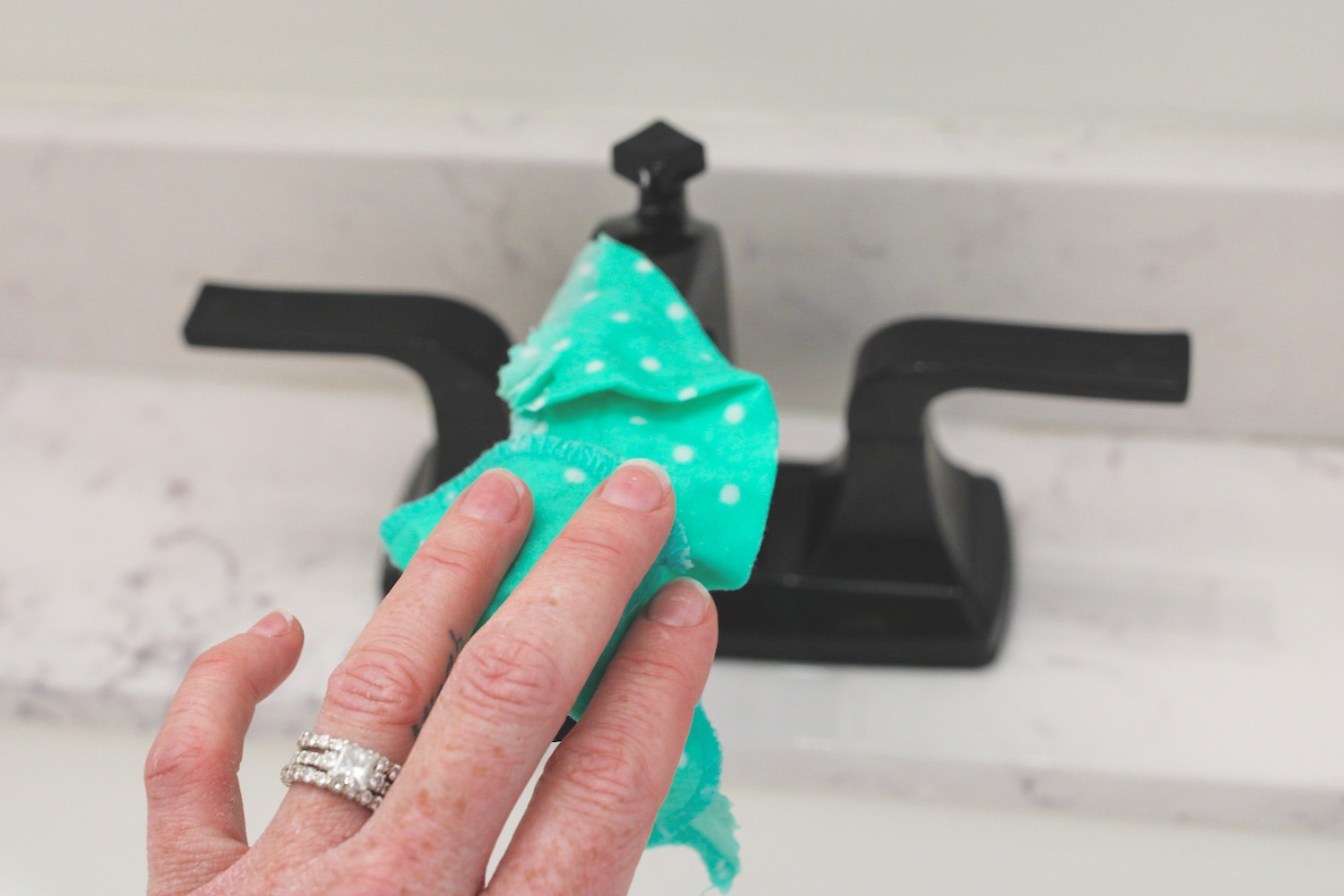 hand wearing diamond wedding bands uses a turquoise cotton cloth to wipe a black bathroom faucet
